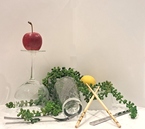 A group of objects on a table including an apple, a wine glass, chopsticks and a glassa plant   