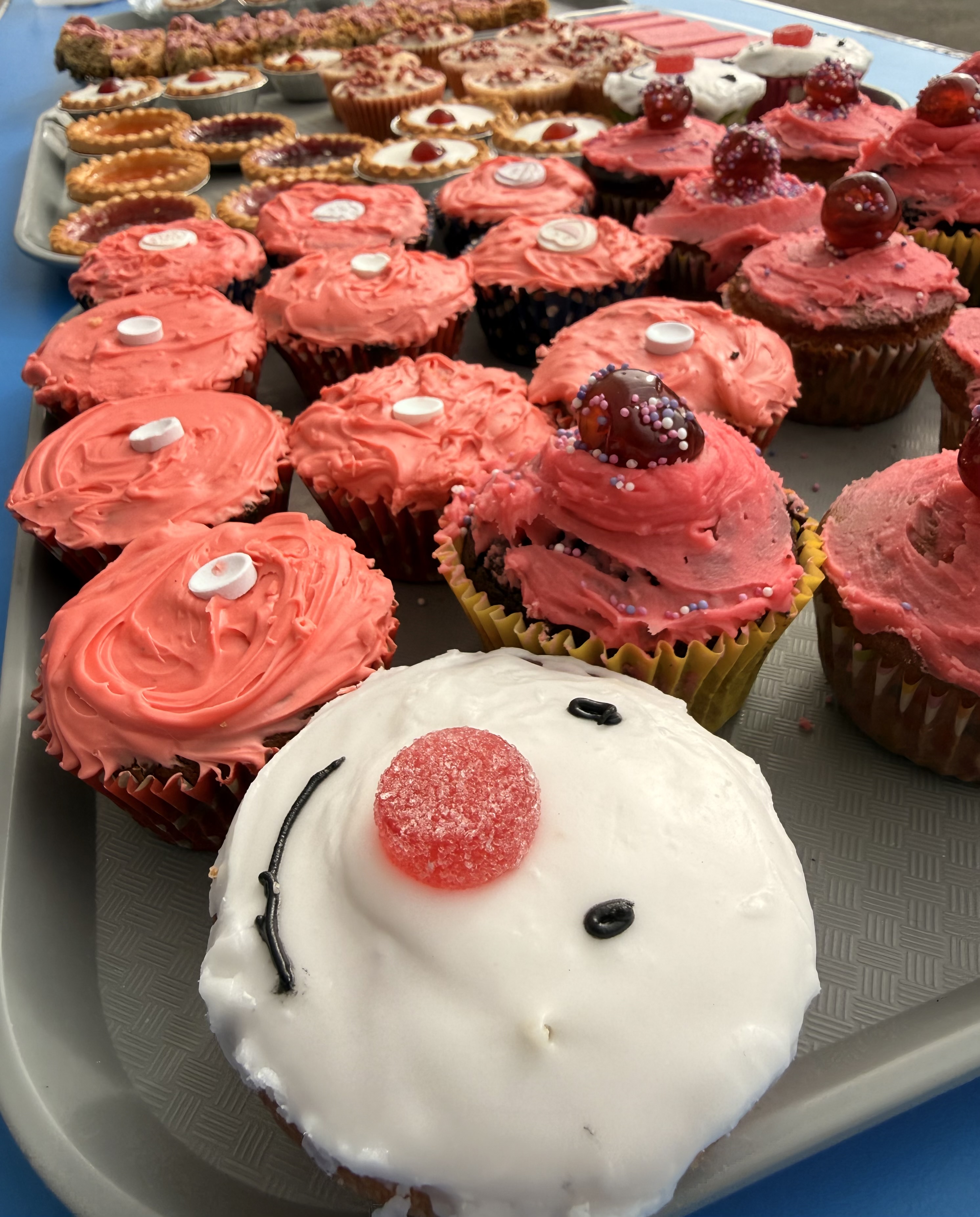 Photograph of a tray filled with various decorated cupcakes and tarts, showcasing a range of pink and red frostings with cherries and candy toppings. A prominent cupcake in the foreground features white icing with a red candy nose and black icing details resembling a face.