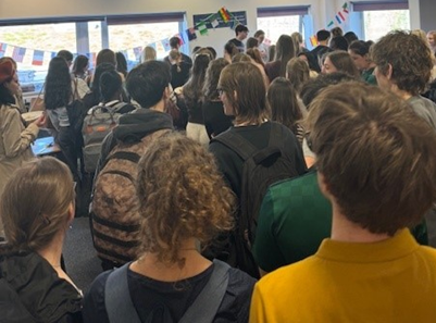 Photograph of a crowded indoor room filled with students, facing towards the front where an event is occurring. The room has white walls, ceiling lights, windows with natural light, and international flags hanging as decoration