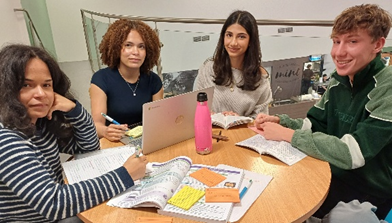 Photograph of four students sitting around a round table engaged in group study, with open notebooks, textbooks, and a laptop visible. Key elements include a pink water bottle at the centre and various study materials spread across the table