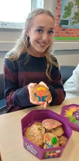 Photograph of a person holding a decorated cookie shaped like a smiling sun with a face, sitting at a table with an open purple tin containing several decorated cookies. Background includes a window and a map on a brick wall,
