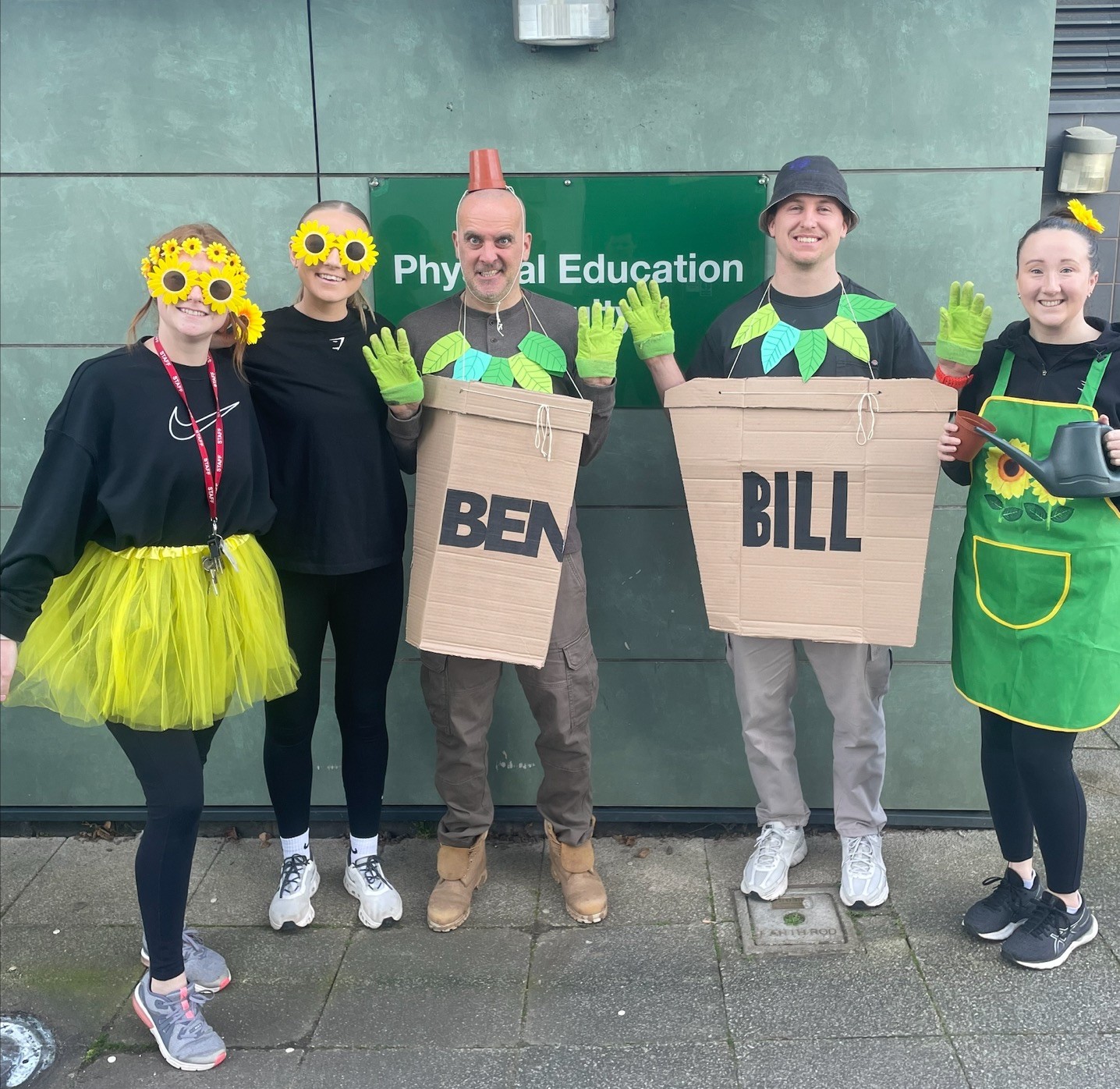 Photograph of five people standing in front of a green wall with a "Physical Education" sign, dressed in colourful costumes including two individuals wearing cardboard signs labelled "BEN" and "BILL." Group features bright yellow, green, and black clothing with accessories of gloves, tutus, and apron.