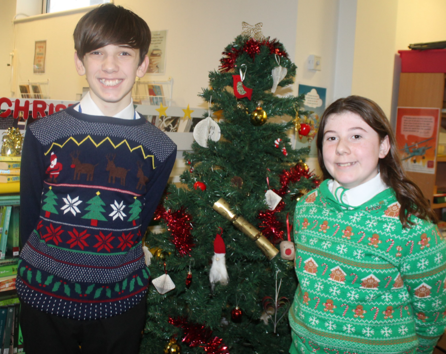 A girl and boy wearing Christmas jumpers standing by a Christmas tree