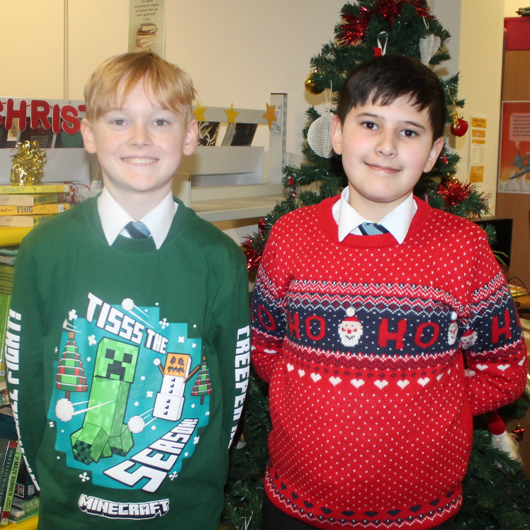 Two boys wearing Christmas jumpers standing in front of a Christmas tree.