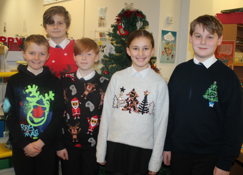 Five students wearing Christmas jumpers by a Christmas tree