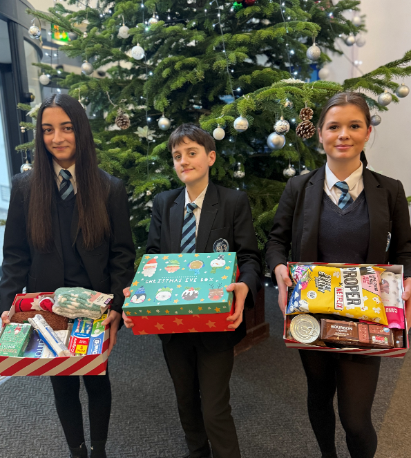 Three students in school uniform holding a Christmas hamper each, standing in front of a Christmas tree