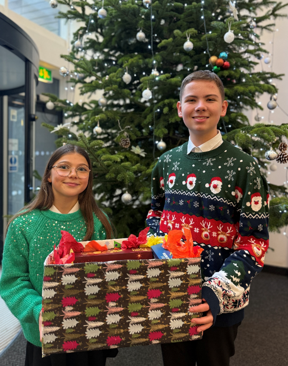 Boy and girl wearing Christmas jumpers holding a Christmas hamper, standing in front of a Christmas tree